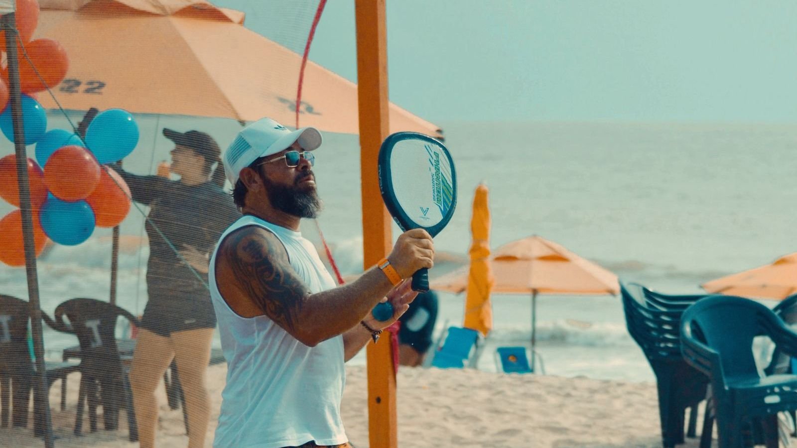 Homem jogando beach tênis na praia, com guarda-sóis ao fundo