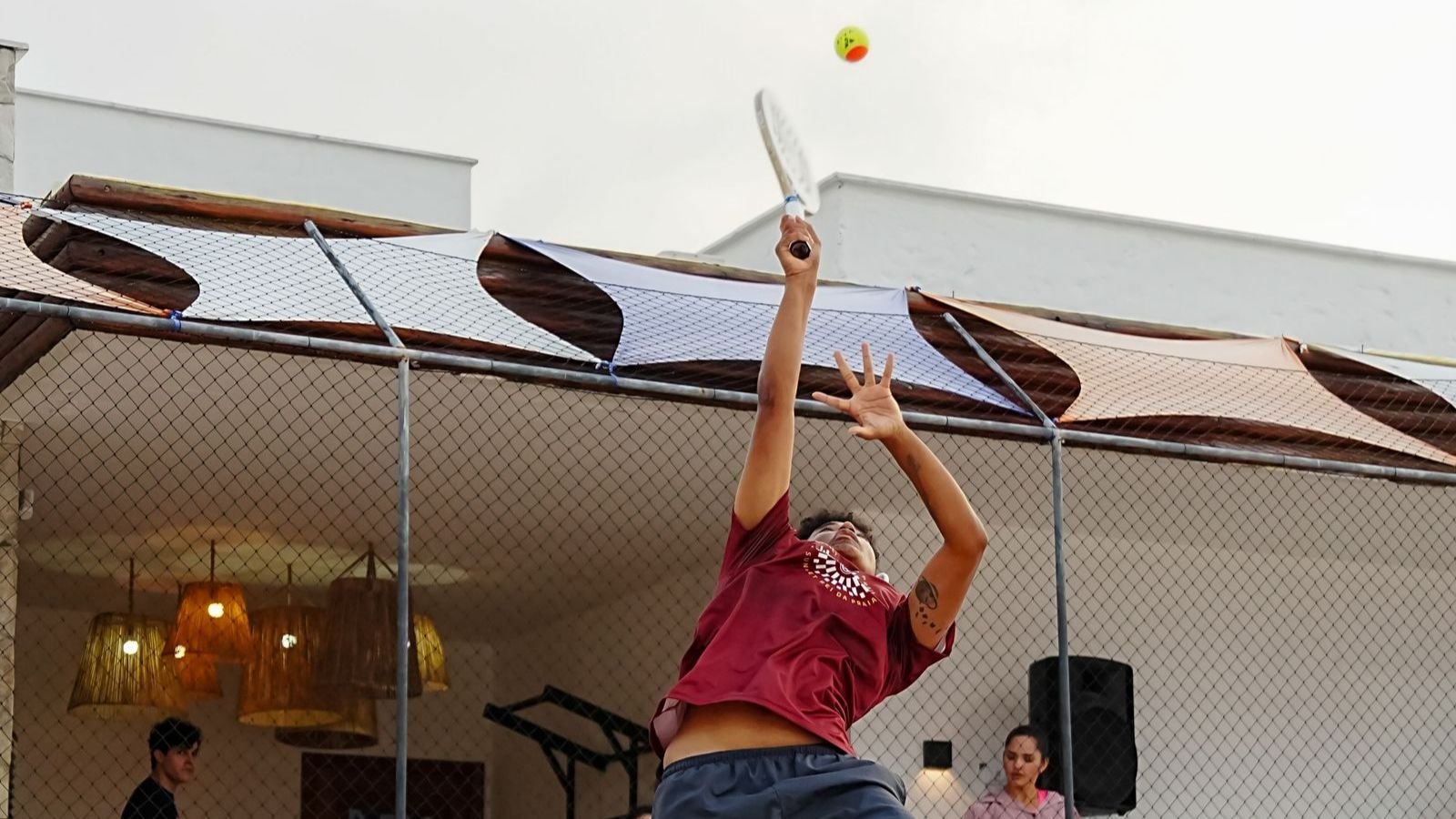 Jogador de beach tênis salta para um smash com a raquete acima da cabeça, bola no alto