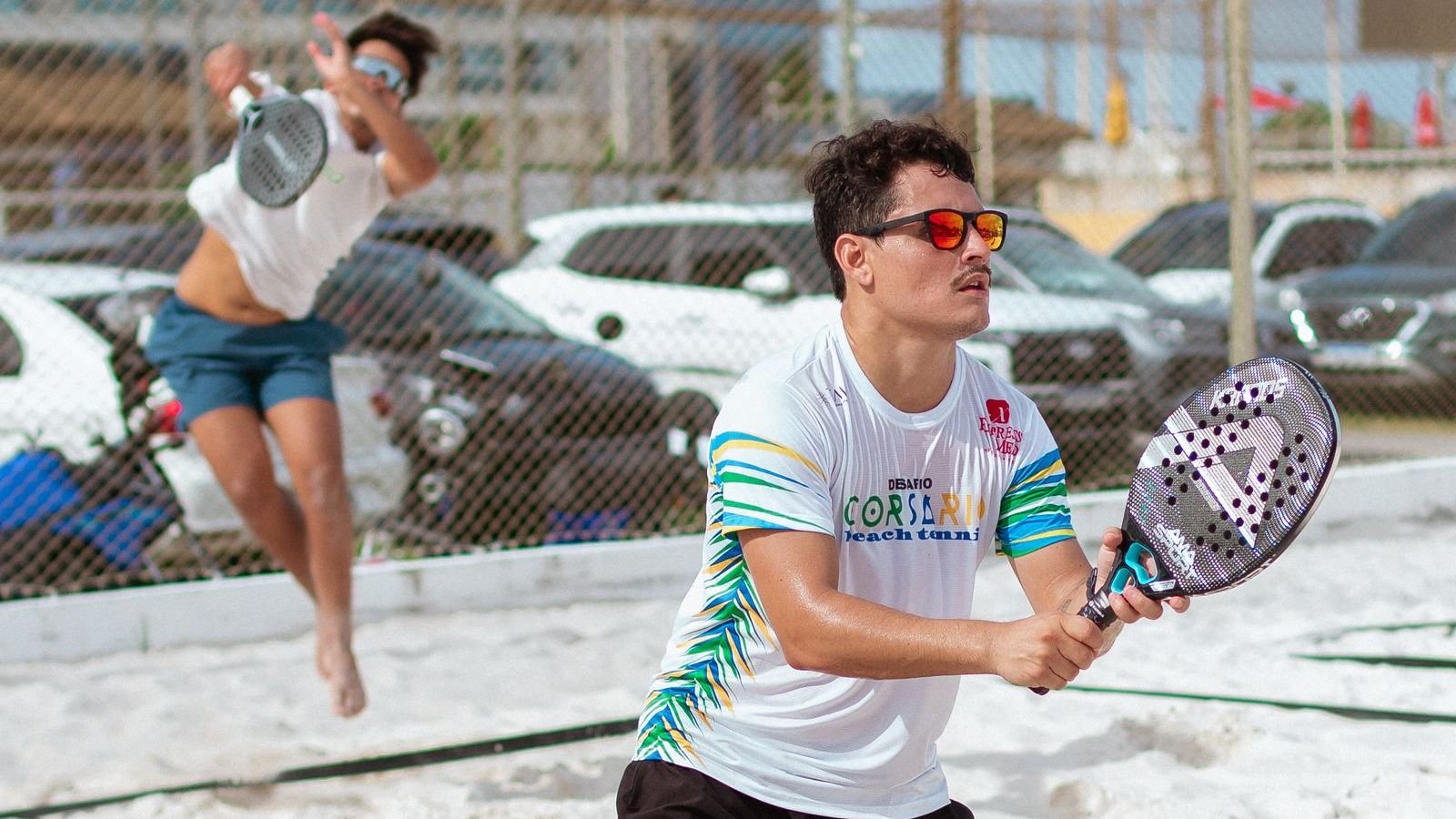 Jogadores de beach tênis de óculos escuros numa quadra de areia em dia de sol forte