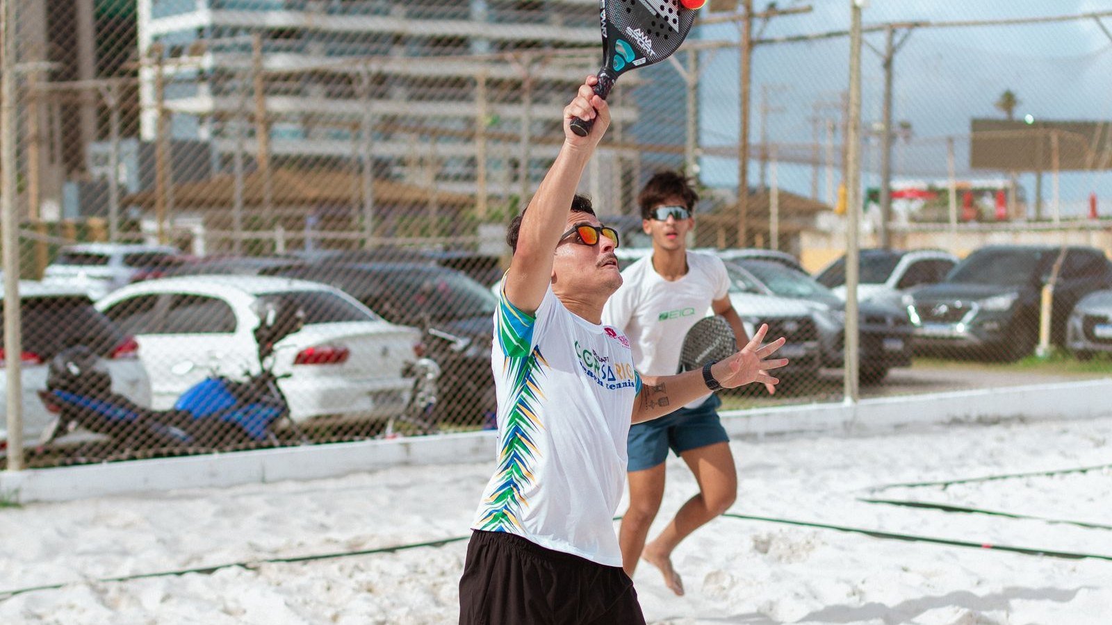 Jogador de beach tênis de óculos escuros esticando o braço acima da cabeça para golpear a bola numa quadra de areia