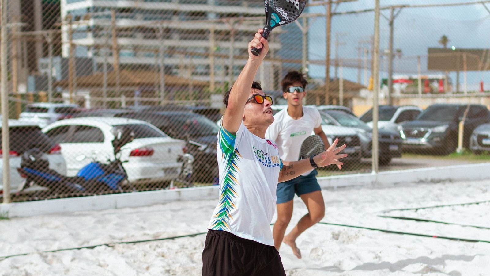 Jogador de beach tênis salta para um smash na quadra de areia, com o parceiro ao fundo